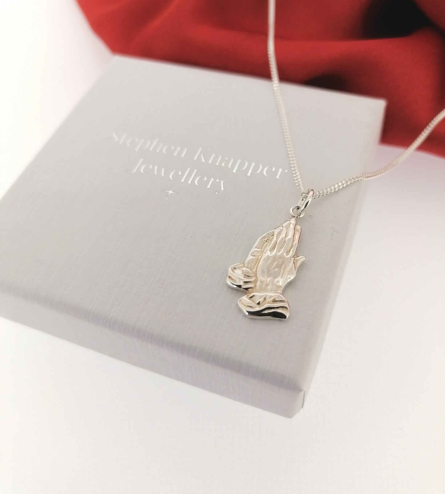 Silver necklace with a praying hands pendant on a Stephen Knapper Jewellery box.