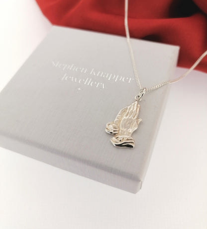 Silver necklace with a praying hands pendant on a Stephen Knapper Jewellery box.